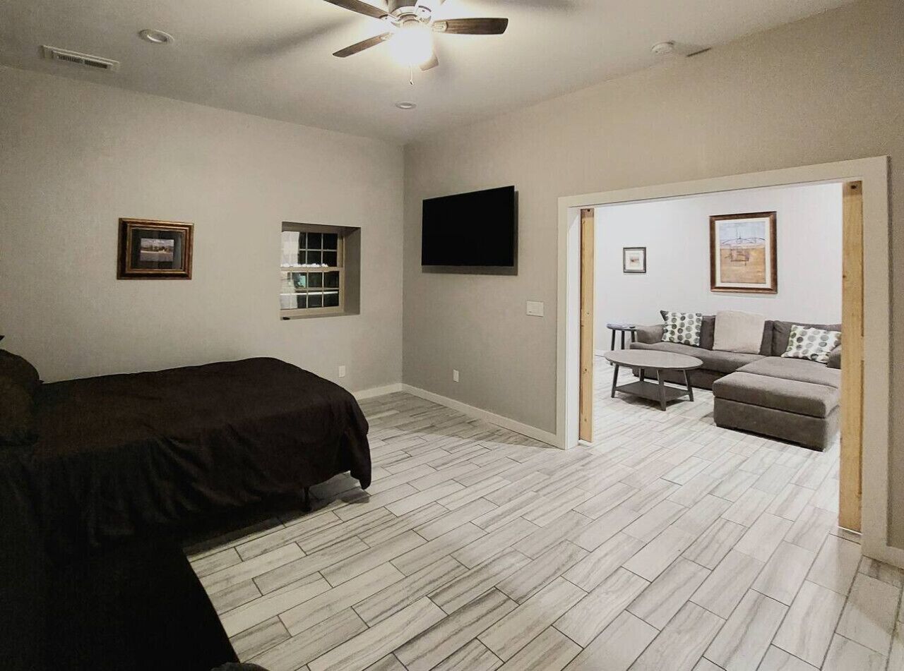 Une chambre avec un lit, un ventilateur de plafond, une télévision et une porte donnant sur un espace de vie comprenant un canapé et une table basse.