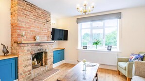 A living room with a brick fireplace, a wooden dining table, a sofa, and a large window with a view of greenery. - 2 bedroom accommodation in Snitterby (Snitterby)