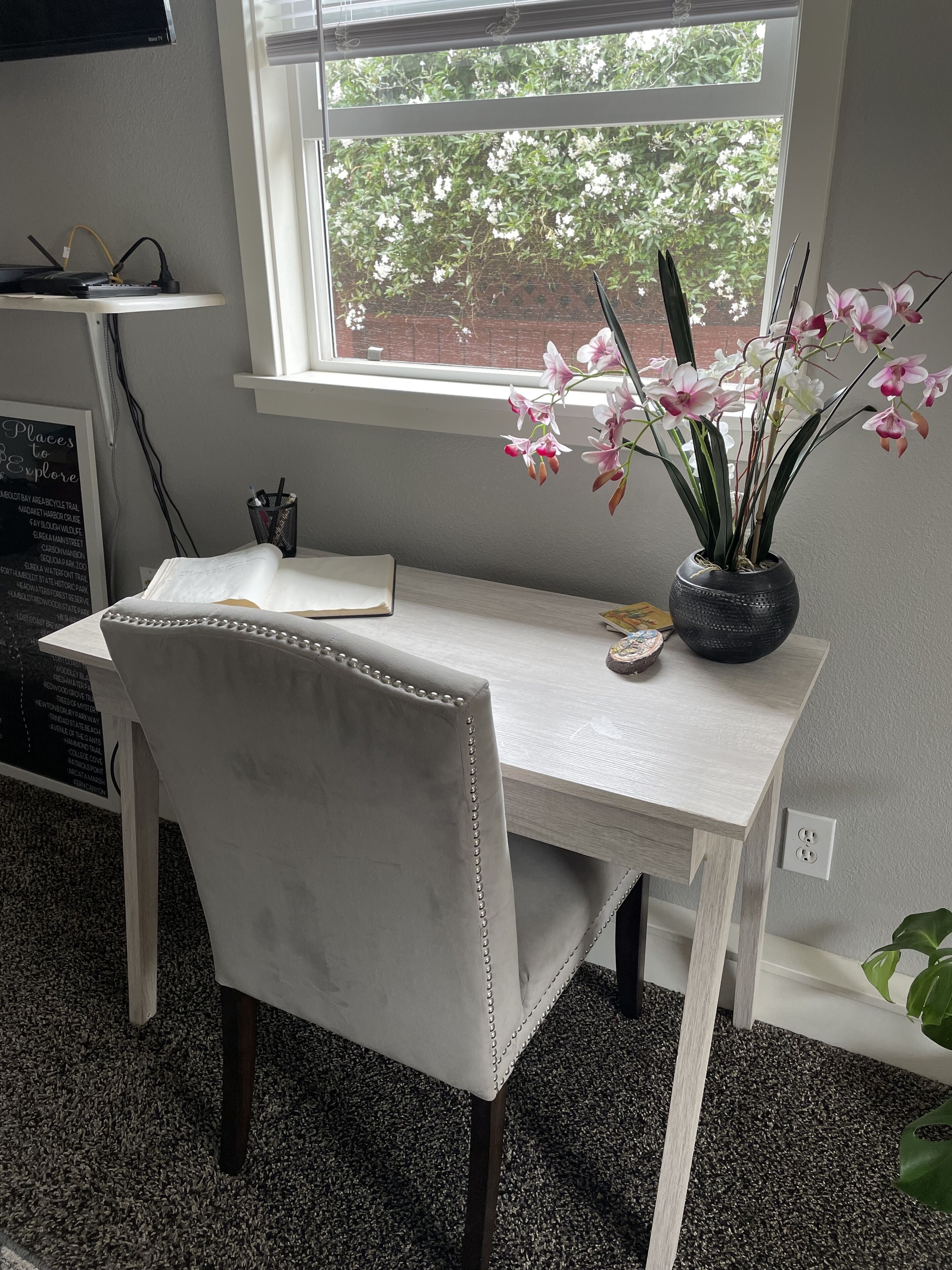 A room with a white desk, a chair, a potted orchid, and a window with a view of trees and flowers.
