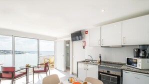 A modern kitchen with a view of the sea, a dining area with a table set for two, and a balcony with red chairs.