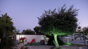 A playground with a slide and swings, surrounded by a white wall and a tree with green lighting.