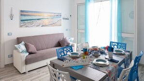 A cozy living room with a sofa, a dining table set for two, and a beach-themed painting on the wall.