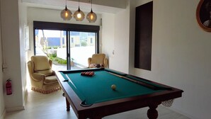 A pool table with a green felt surface, a pool cue, and a cue ball. There is a beige armchair and a red fire extinguisher on the wall. - The Villa Reina (Le Croisic)