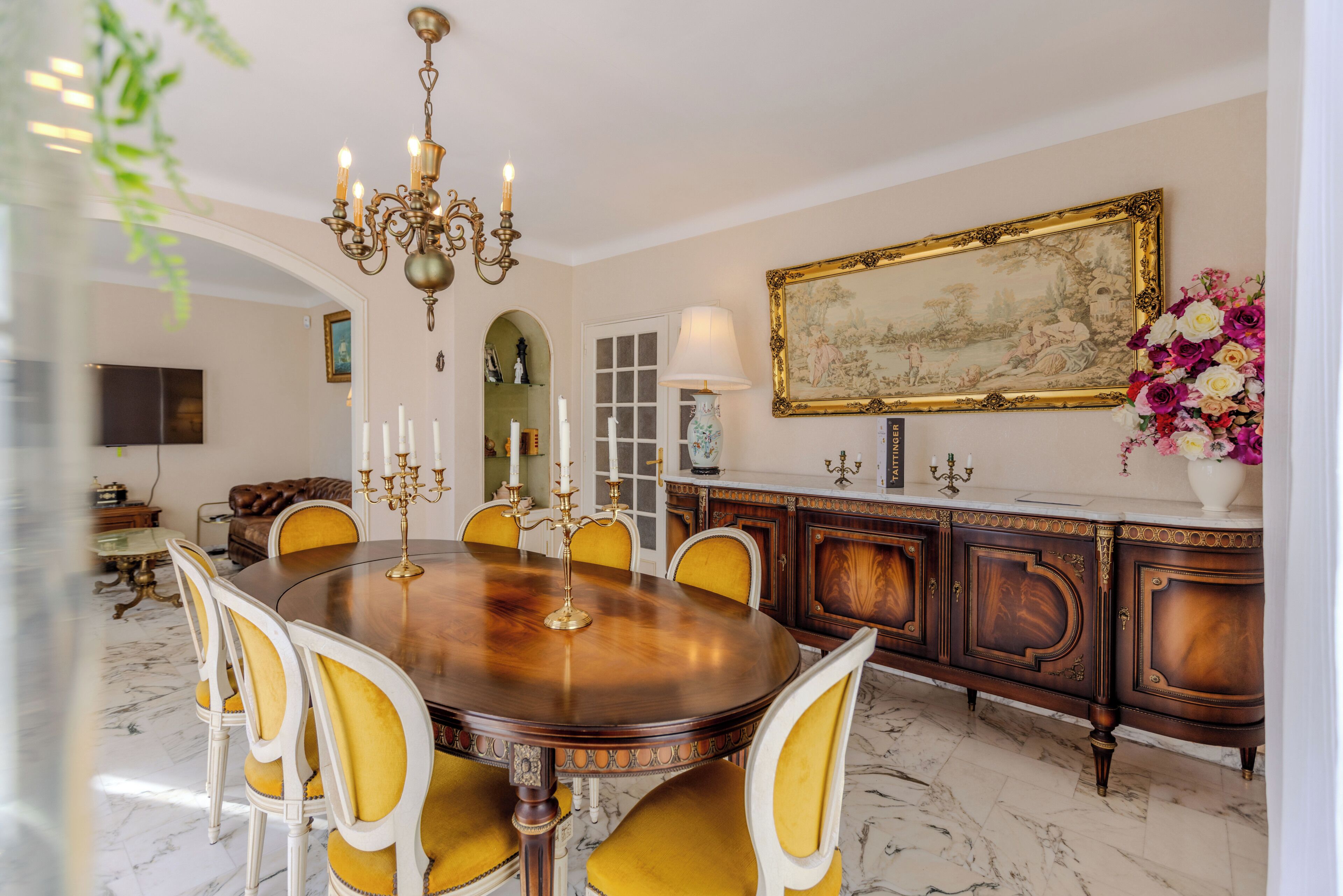 Una sala da pranzo con un tavolo rotondo, sedie, un lampadario, un grande quadro e un mobile con un vaso di fiori.