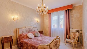 A bedroom with a large bed, a chair, a nightstand, a chandelier, and a window with curtains. - The Villa Reina (Le Croisic)