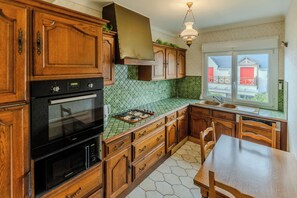 A kitchen with wooden cabinets, a gas stove, a built-in oven, and a window with a view of a house. - The Villa Reina (Le Croisic)