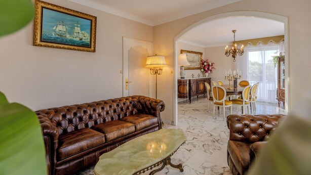 A living room with a brown leather sofa, a matching armchair, a marble coffee table, a dining area with a chandelier, and a painting of ships on the wall. - The Villa Reina (Le Croisic)