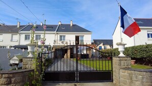 A gated residential area with a flag, a paved driveway, and a house with a balcony. - The Villa Reina (Le Croisic)