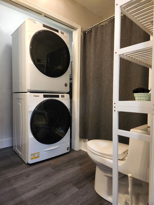 A laundry room with two stacked Whirlpool washers and dryers, a toilet, and a shelving unit with a basket.