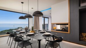A modern dining room with a panoramic sea view, a large dining table set for six, and a built-in fireplace.