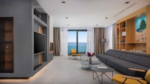 A modern living room with a grey sofa, yellow chairs, a flat-screen TV, and a view of the sea through large windows.