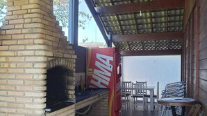 An outdoor patio with a brick barbecue grill, a table with chairs, and a covered area with a corrugated metal roof. - Heated pool **FREE** + Air conditioning + 300MB Vivo fiber internet, Riviera Beach (Bertioga)