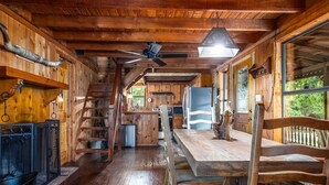 A rustic cabin interior with wooden walls, a dining area, a ceiling fan, and a large window. - Old Fellow's Farmhouse (Kerrville)