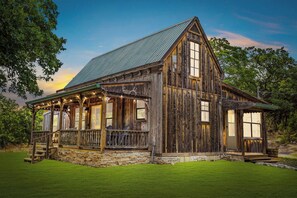 A rustic wooden building with a stone foundation, large windows, and a porch with railings. - Old Fellows Farmhouse | 2BR with Country Views (Kerrville)