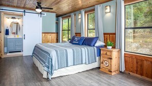 A bedroom with a large bed, a wooden nightstand, a mirror, and a window with curtains. - Old Fellow's Farmhouse (Kerrville)