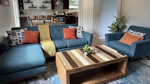 A modern living room with a sectional sofa, a coffee table, and a kitchen in the background. - 3 KING BEDS - 3 QUEEN - 4 TWINS - 5 BED ROOMS (Houston)