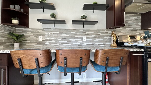 A modern kitchen with a tiled backsplash, wooden bar stools, and built-in shelves with plants. - 3 KING BEDS - 3 QUEEN - 4 TWINS - 5 BED ROOMS (Houston)