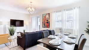Un salon moderne avec un canapé foncé, une table à manger dressée pour quatre personnes et un grand téléviseur fixé au mur.