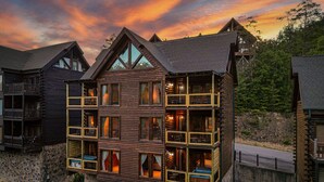 A multi-story log cabin with balconies and large windows, surrounded by a stone wall and set against a forest backdrop.