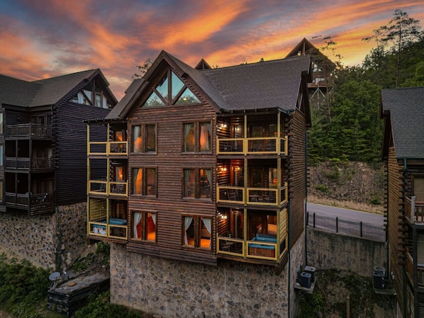 A multi-story log cabin with balconies and large windows, surrounded by a stone wall and set against a forest backdrop.