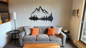 A modern living room with a grey sofa, two orange pillows, a wooden coffee table, a black floor lamp, and a wall art of a mountain landscape. - Smooth 2-minute ski to Lift Ski-in Ski-out • Smuggs Condo ⭐️  (Jeffersonville)