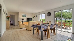 A modern living room with a sofa, a dining table set for four, and a TV. There is a large window providing a view of the outdoors. - Le Clos des Pommiers (PAIMPOL)