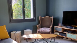 A living room with a sofa, armchair, coffee table, TV stand, and a window with a view of greenery. - Charming seaside vacation home (Pénestin)