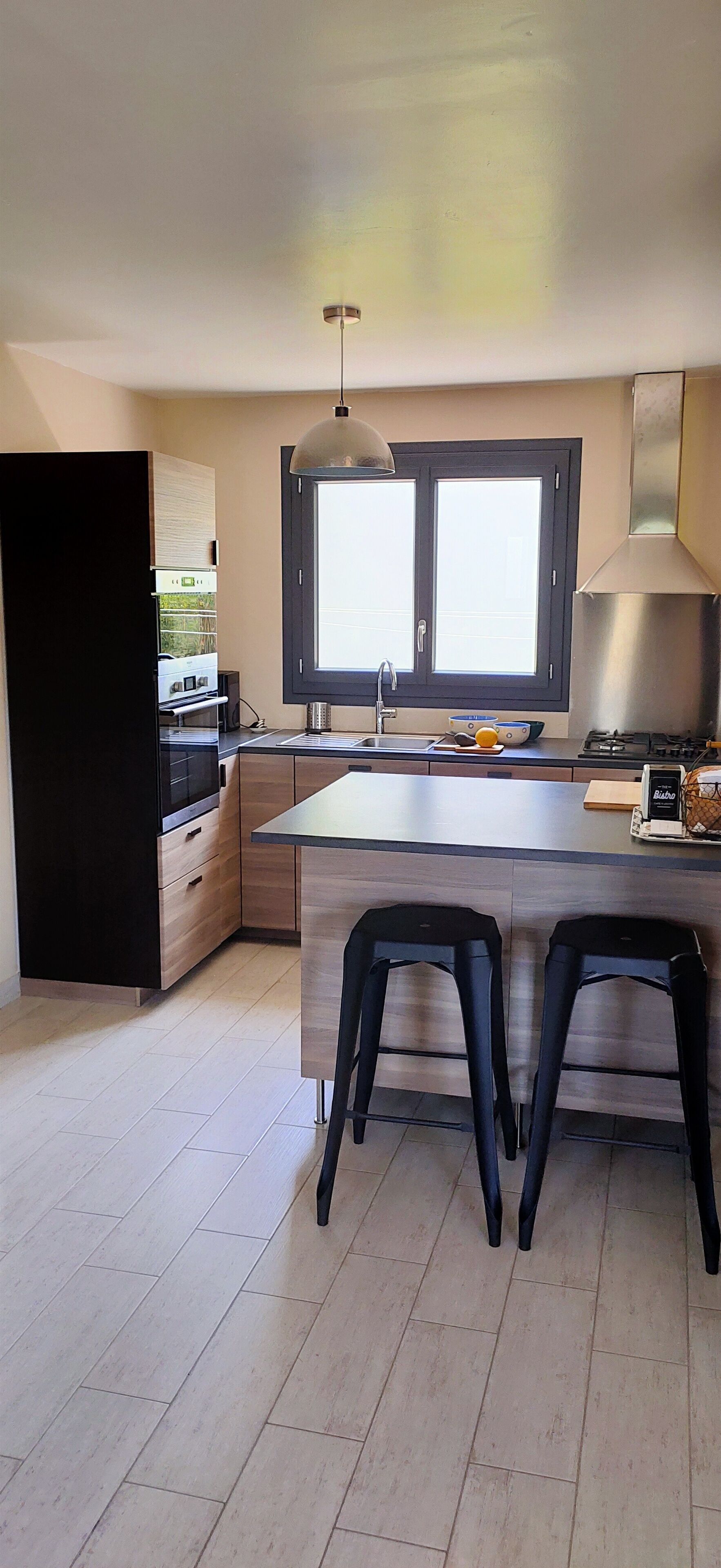 A modern kitchen with a central island, black bar stools, a window with blinds, and a stainless steel range hood.