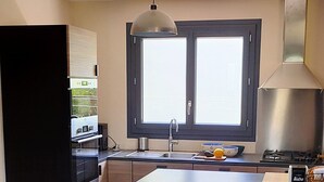 A modern kitchen with a central island, black bar stools, a window with blinds, and a stainless steel range hood. - Charming seaside vacation home (Pénestin)