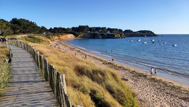 A wooden boardwalk leading to a sandy beach with clear blue water, surrounded by grassy dunes and a few houses in the distance. - Charming seaside vacation home (Pénestin)