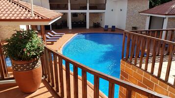 A swimming pool with a wooden deck and lounge chairs, surrounded by a tiled deck and a brick wall.