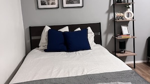 Ein ordentlich bezogenes Bett mit einer weiß-grauen Bettdecke, zwei blauen Kissen und einer grauen Tagesdecke. Darüber an der Wand hängen zwei gerahmte Bilder, darunter ein schwarzes Metallregal mit Dekoration und ein kleiner Tisch mit einer Lampe.