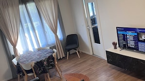 Ein Wohnzimmer mit einem schwarzen Ledersofa, einem kleinen Holztisch mit Fernbedienung, einem Esstisch mit Stühlen und einem Fernseher auf einem Lowboard.