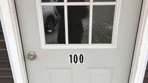 Een witte deur met het nummer 100, een deurmat waarop 'WELKOM' staat, en een raam dat een persoon en een fiets reflecteert.