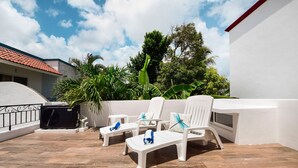 A rooftop terrace with white lounge chairs, a small table, and a black grill.