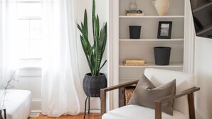 Een woonkamer met een witte fauteuil, een zwarte vloerlamp, een plant in pot en een witte boekenkast met decoratieve spullen.