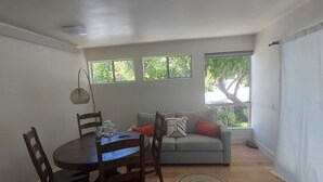 A dining area with a round table, chairs, and a lamp. There is a sofa with cushions and a window with a view of greenery. - Attached Unit, a few minute walk from the Beach (Santa Barbara)