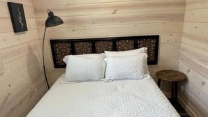 Uma cama com colcha branca, luminária preta e uma pequena mesa redonda num quarto com paredes de madeira.