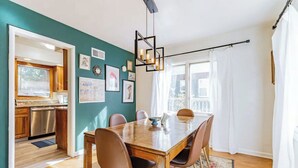 Un comedor con una mesa de madera, cuatro sillas y una araña. Se ve una cocina a través de un vano abierto.