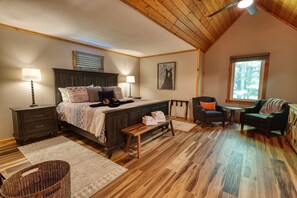 A bedroom with a wooden ceiling, a bed with patterned bedding, two bedside lamps, a chair, a small table, and a window with a view of trees.