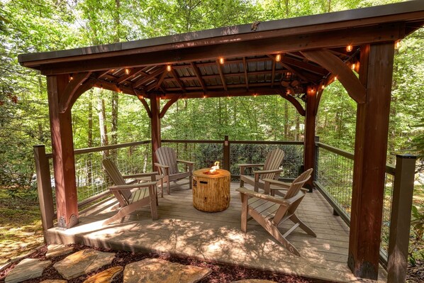 A wooden gazebo with a fire pit and four Adirondack chairs.