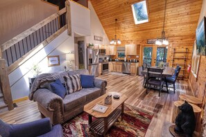 Un accogliente interno di una baita con una scala in legno, una zona giorno con divano e tavolino, una cucina con elettrodomestici moderni e un'area pranzo con tavolo e sedie in legno.
