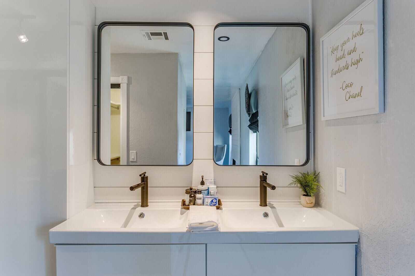A bathroom with a white vanity, two sinks, and a large mirror. There is a framed quote on the wall and a small plant on the vanity.