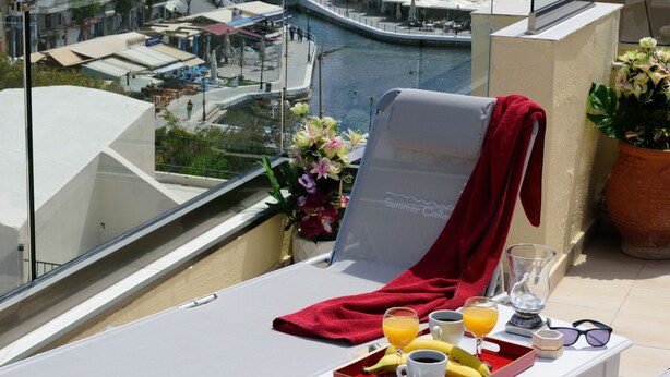 A balcony with a lounge chair, a tray with breakfast items, and a view of a coastal town with buildings and a marina.