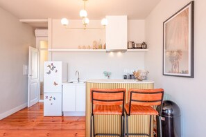 Una cocina compacta con armarios blancos, un lavamanos y una encimera. Hay dos sillas naranjas con estructura negra y un cubo de basura negro.