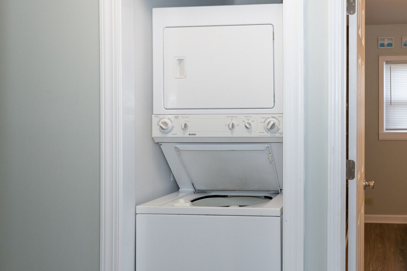 A compact, white, wall-mounted washer and dryer unit in a small laundry room with a wooden door and a window with blinds.