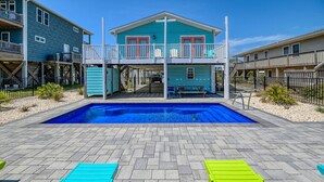 Een zwembadgedeelte met een turquoise huis, een houten terras en twee bankjes.