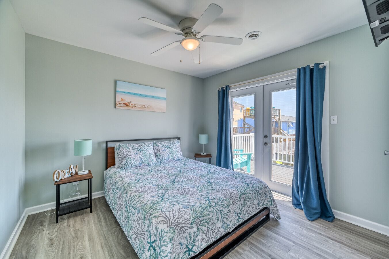 A bedroom with a bed, a ceiling fan, a nightstand, a lamp, a painting, and a view of the outside.