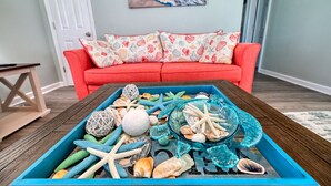 Een kamer met een blauwe dienblad vol met schelpen en zeesterren, een rode bank met gestreepte kussens, en een schilderij van een zandstrand aan de muur.
