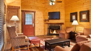A cozy log cabin interior with a fireplace, a television mounted above it, and a comfortable seating area with a patterned armchair and ottoman.